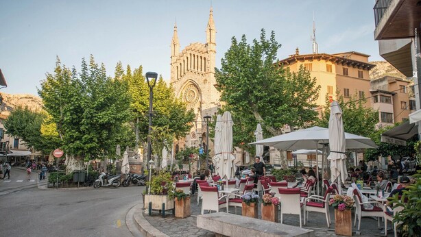 An outdoor cafe with tables and chairs, surrounded by trees and buildings, including a prominent church in the background.