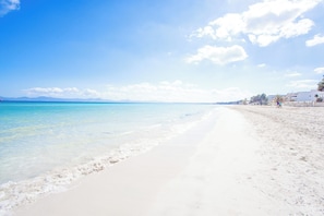 A beach with clear turquoise water, white sand, and a few buildings in the background.