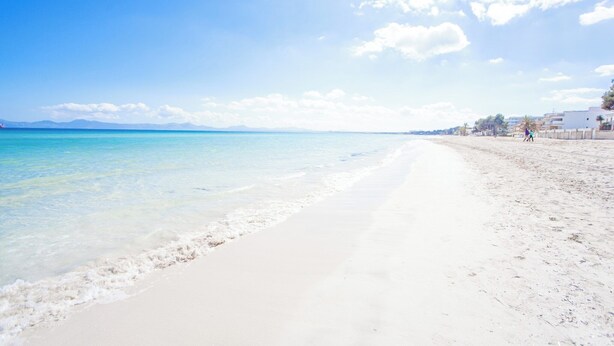 A beach with clear turquoise water, white sand, and a few buildings in the background. - Sun Of The Bay Vidalba 2 (b2 - A2) - Apartment (Port D'alcudia, Illes Balears)