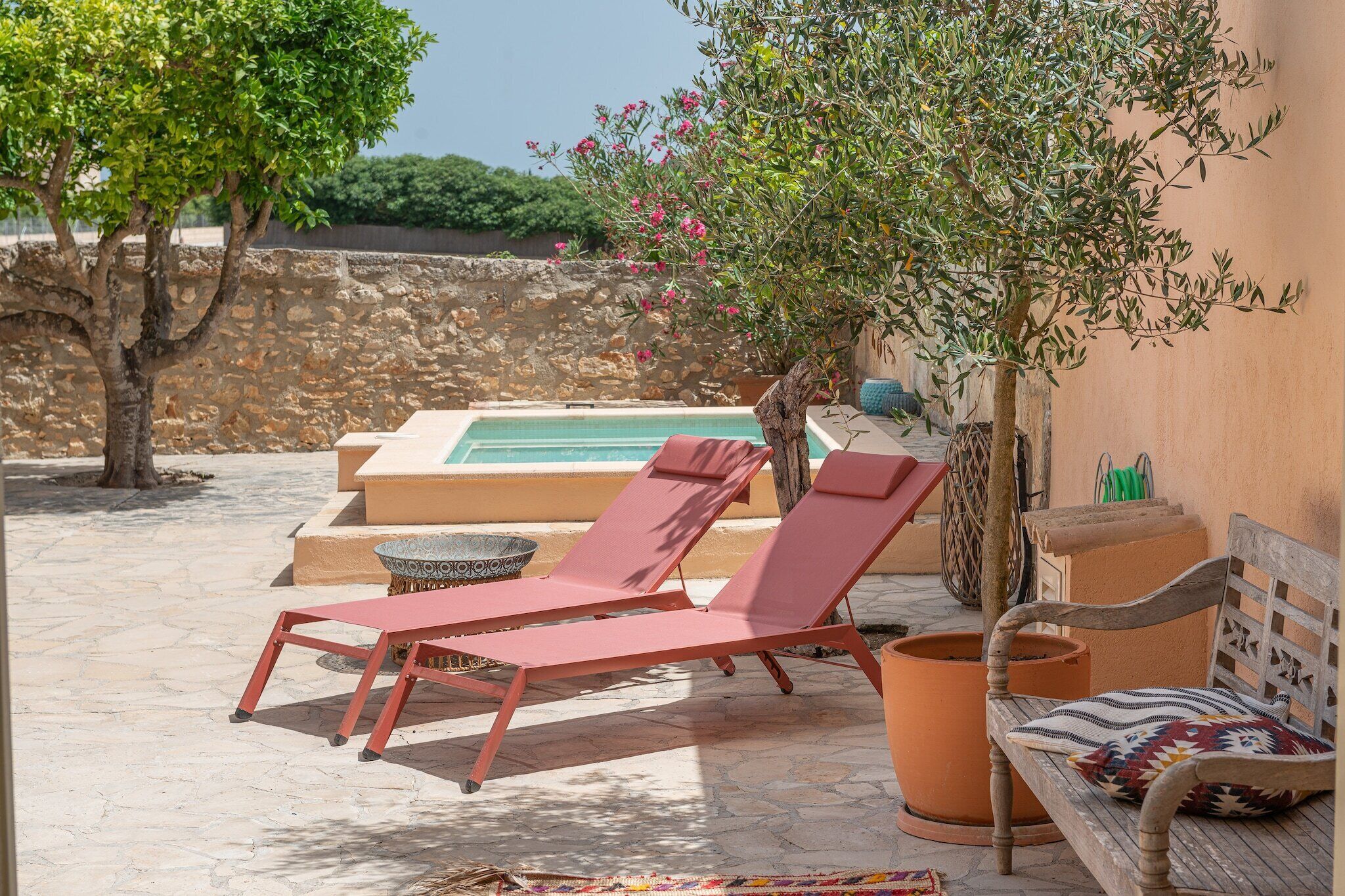 Zona de piscina con sillas de descanso, un banco de madera y una pared de piedra al fondo.
