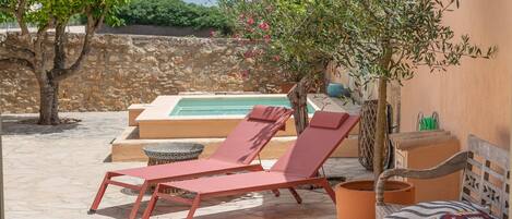 Zona de piscina con sillas de descanso, un banco de madera y una pared de piedra al fondo.
