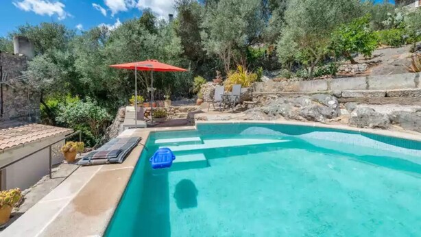 Una piscina con un paraguas rojo, sillas de descanso y un muro de piedra.
