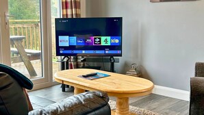 A living room with a TV, a wooden table, a sofa, and a view of the outdoors. - Skye -  a chalet that sleeps 3 guests  in 2 bedrooms (Oban)