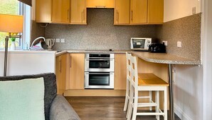 A compact kitchen with wooden cabinets, a built-in oven, a microwave, and a small dining area with a white chair. - Skye -  a chalet that sleeps 3 guests  in 2 bedrooms (Oban)