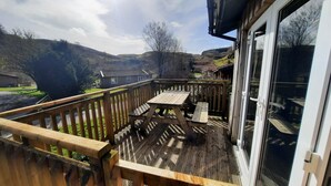A wooden deck with a picnic table, a glass door, and a view of a rural landscape with hills and a house. - Skye -  a chalet that sleeps 3 guests  in 2 bedrooms (Oban)