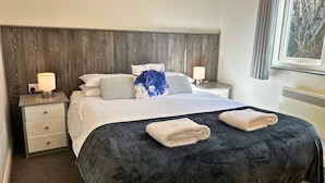 Una cama bien hecha con un edredón oscuro y dos toallas dobladas, un cabecero de madera, ropa de cama blanca y dos lámparas de noche.