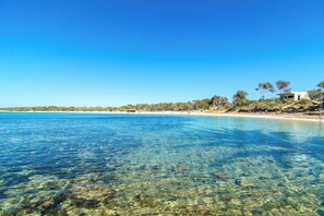 Uma praia costeira com águas claras, areal arenoso e um edifício com fachada branca.