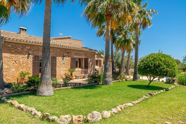 Una casa de piedra con tejado de tejas, rodeada de un césped cuidado y palmeras.