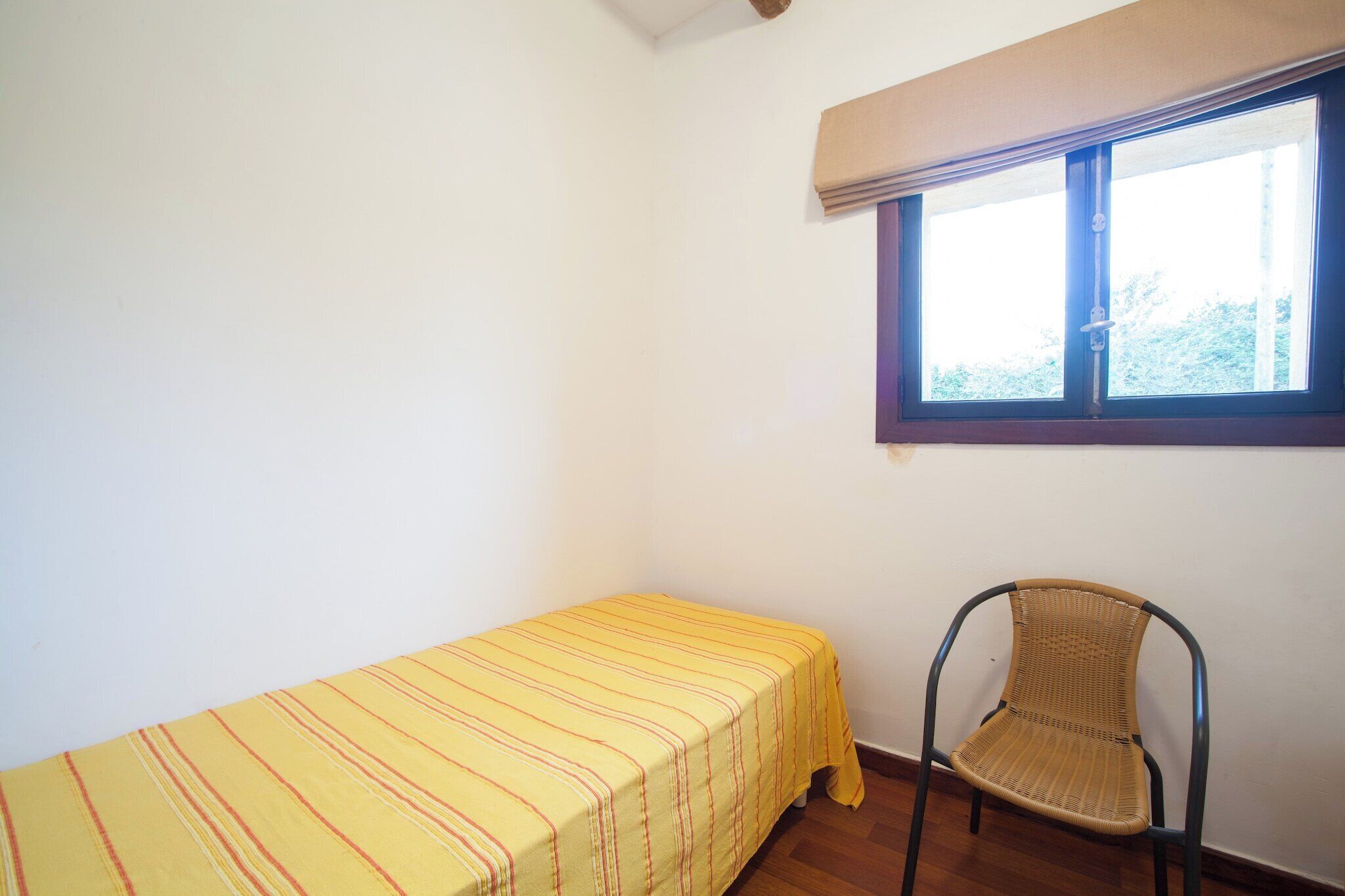 A small, single-bed room with a plain wall, a window with a blind, and a chair.