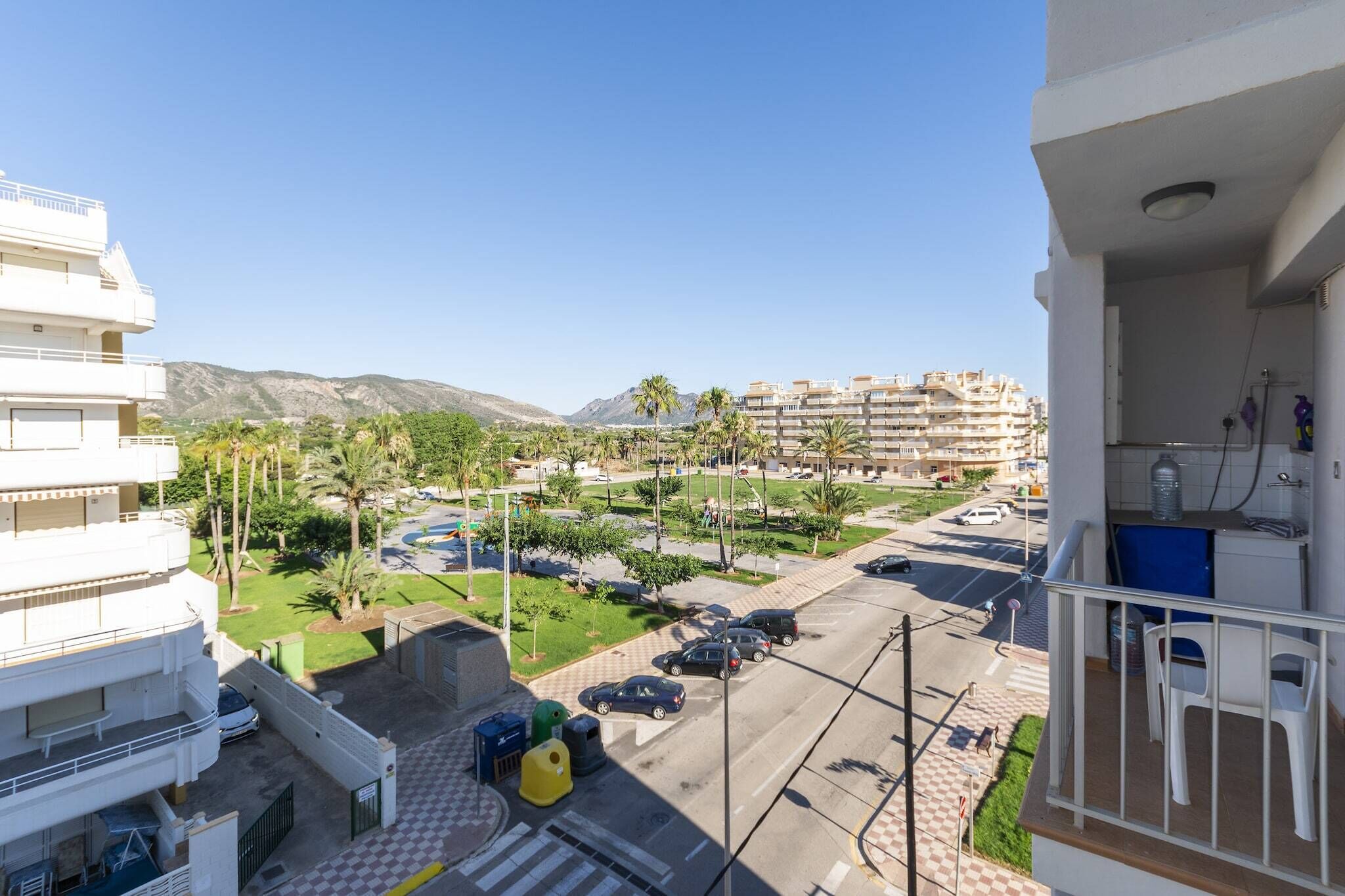 Un edificio residencial con balcón, un estacionamiento con coches y una vista de un parque verde y montañas al fondo.