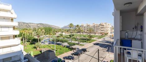 Un edificio residencial con balcón, un estacionamiento con coches y una vista de un parque verde y montañas al fondo.