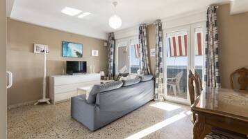 Una sala de estar con sofá, televisión, cómoda blanca, mesa de comedor y balcón con cortinas.