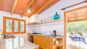 A kitchen with granite countertops, wooden cabinets, a built-in microwave, and a coffee maker. There is a window with a view of trees and a dining area with a table and chairs. - Casa Mas - Villa With Private Pool In Felanitx (Felanitx, Illes Balears)