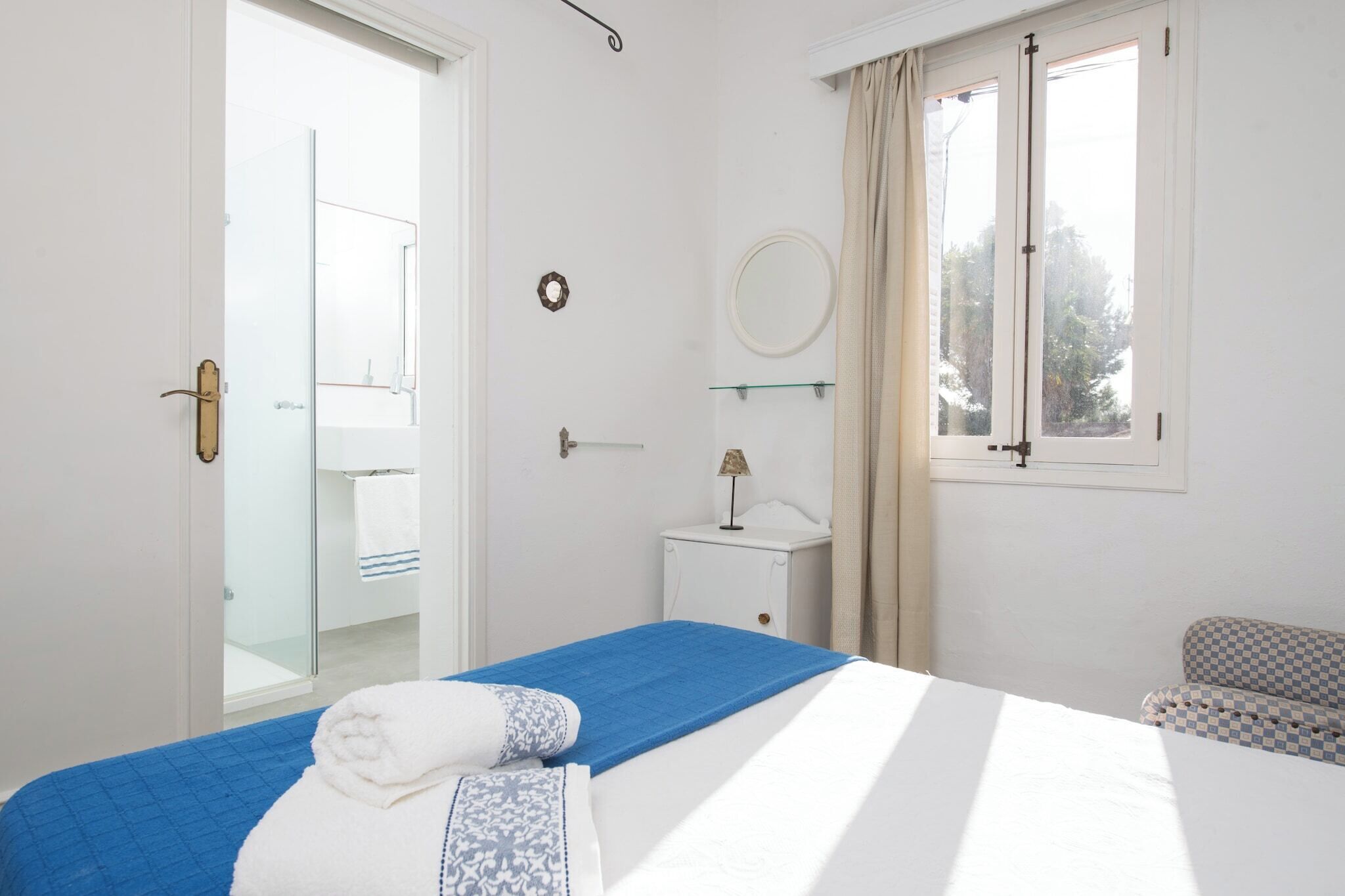 Quarto com cama, mesa de cabeceira, espelho, janela com cortinas e banheiro visível através de uma porta aberta.