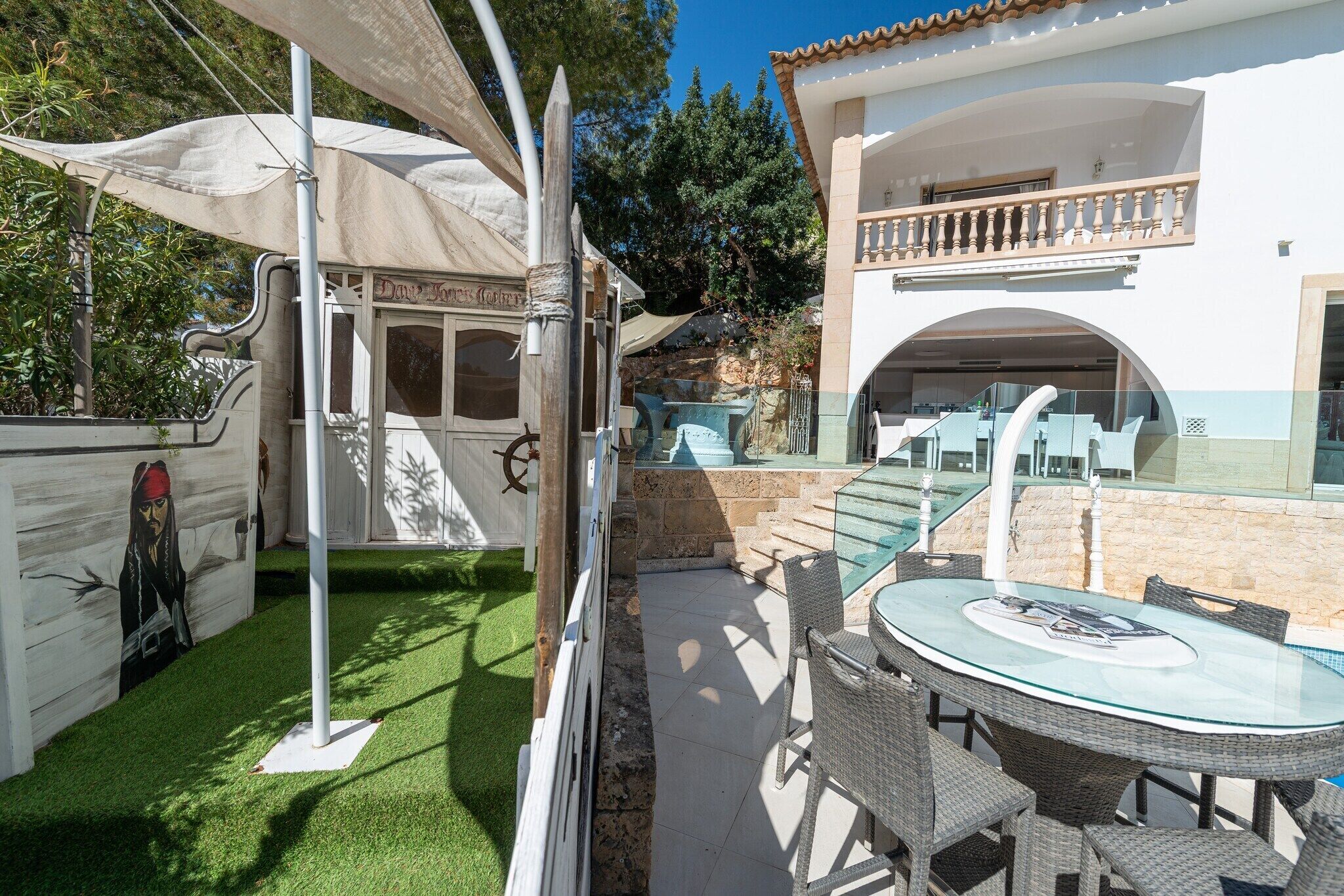 A patio with a glass table and wicker chairs, a white building with a balcony, and a mural on the wall.