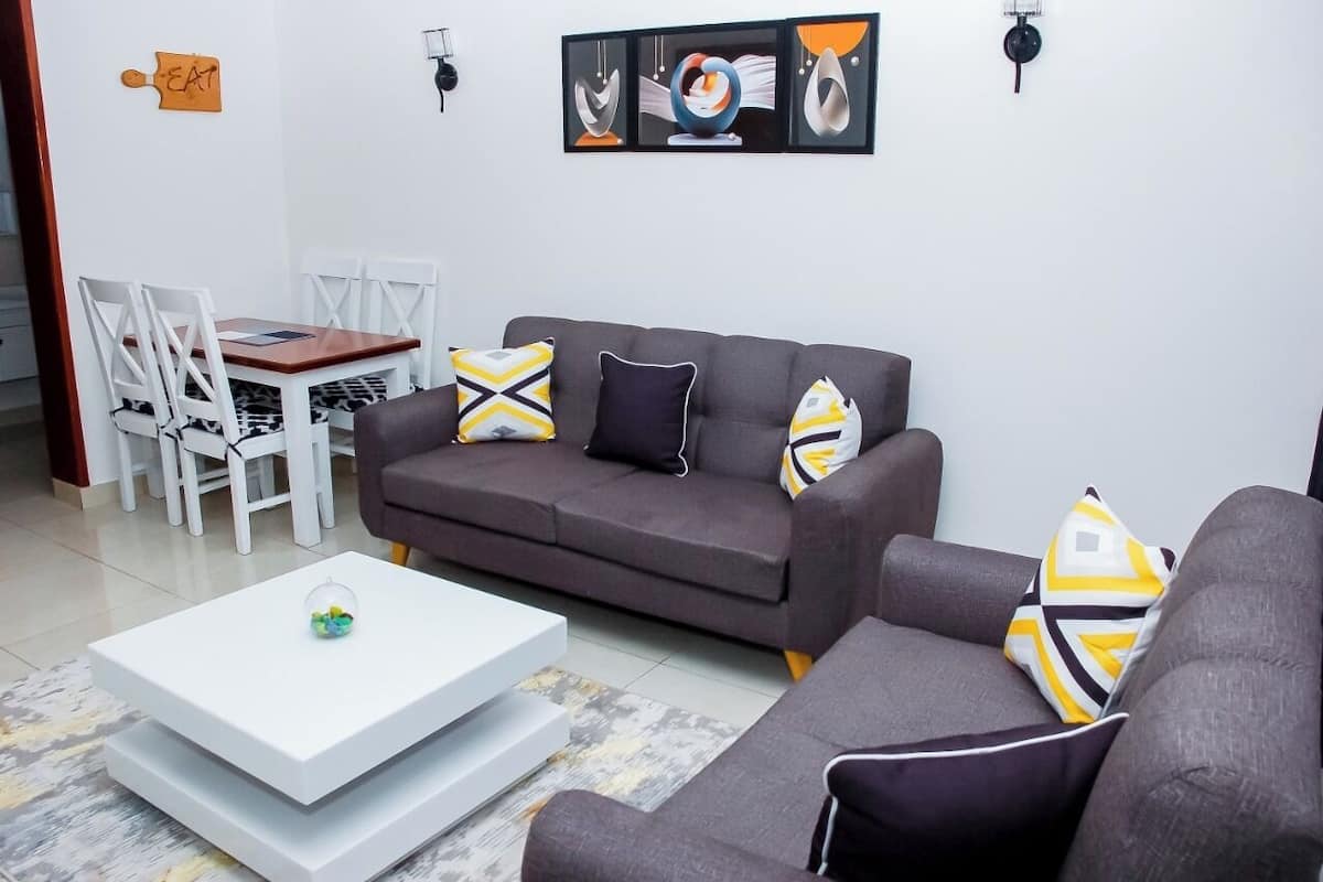A living room with a grey sofa, white coffee table, and patterned cushions. There is a dining table with white chairs and a sign that says "EAT".