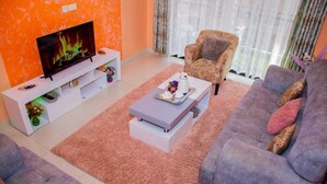 Una sala de estar con un televisor de pantalla plana sobre una consola blanca, un sillón estampado, un sofá gris y una alfombra rosa.