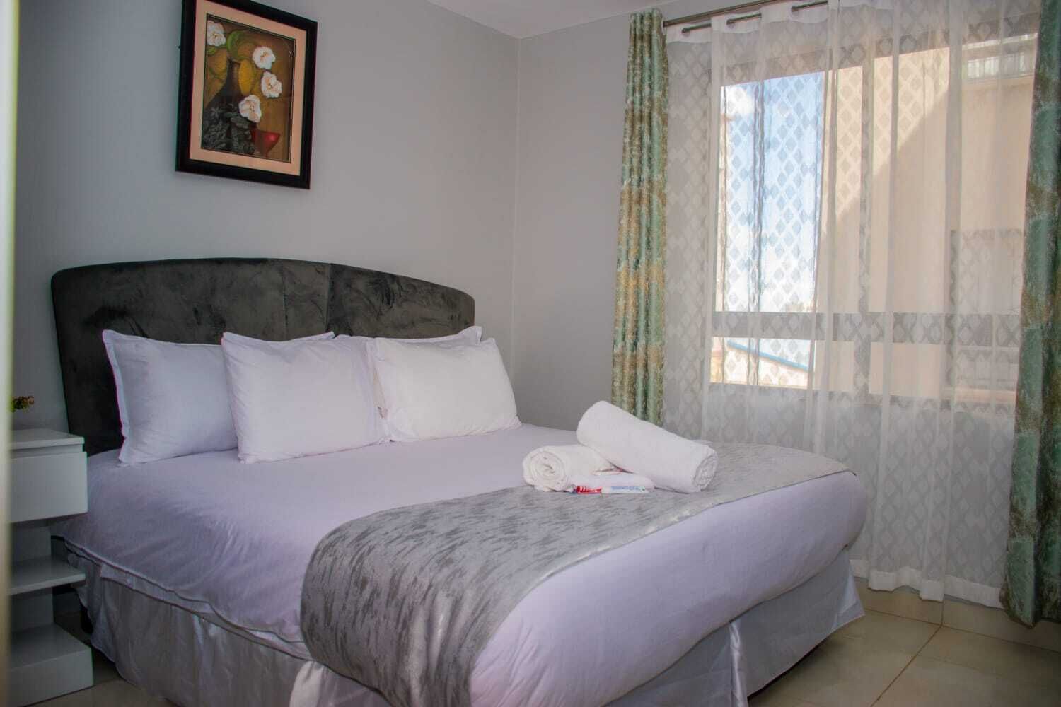A neatly made bed with white linens and pillows, a framed picture on the wall, and a window with sheer curtains.