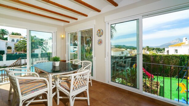 A balcony with a glass door, white wicker furniture, and a view of a playground and mountains. - Gladiolo - Villa With Private Pool (El Tosalet - Xàbia, Alicante)