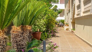 A narrow, paved pathway flanked by lush greenery and potted plants, leading to a building with a tiled roof and balconies. - Llepol - Apartment In Port D'alcúdia. Free Wifi (Port D'alcÚdia, Illes Balears)