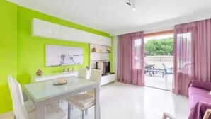 A modern living room with a dining area, a large window with curtains, and a view of greenery outside. - Llepol - Apartment In Port D'alcúdia. Free Wifi (Port D'alcÚdia, Illes Balears)