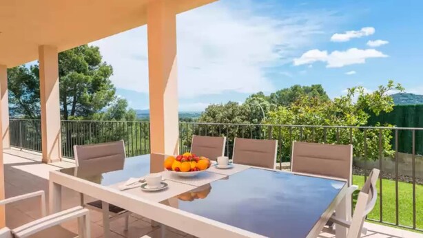 Un espace extérieur abrité, aménagé pour les repas, avec une table et des chaises, un saladier rempli de fruits, et une vue sur la végétation et les collines environnantes.