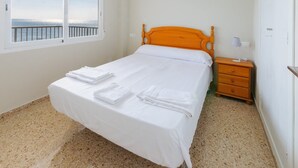 A neatly made bed with white linens and towels, a wooden headboard, a bedside table with a lamp, and a window with a view of the sea.