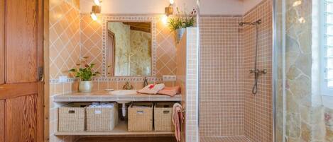 A bathroom with a stone wall, wooden door, tiled floor, a sink with a mirror, a shower, and wicker baskets under the sink.