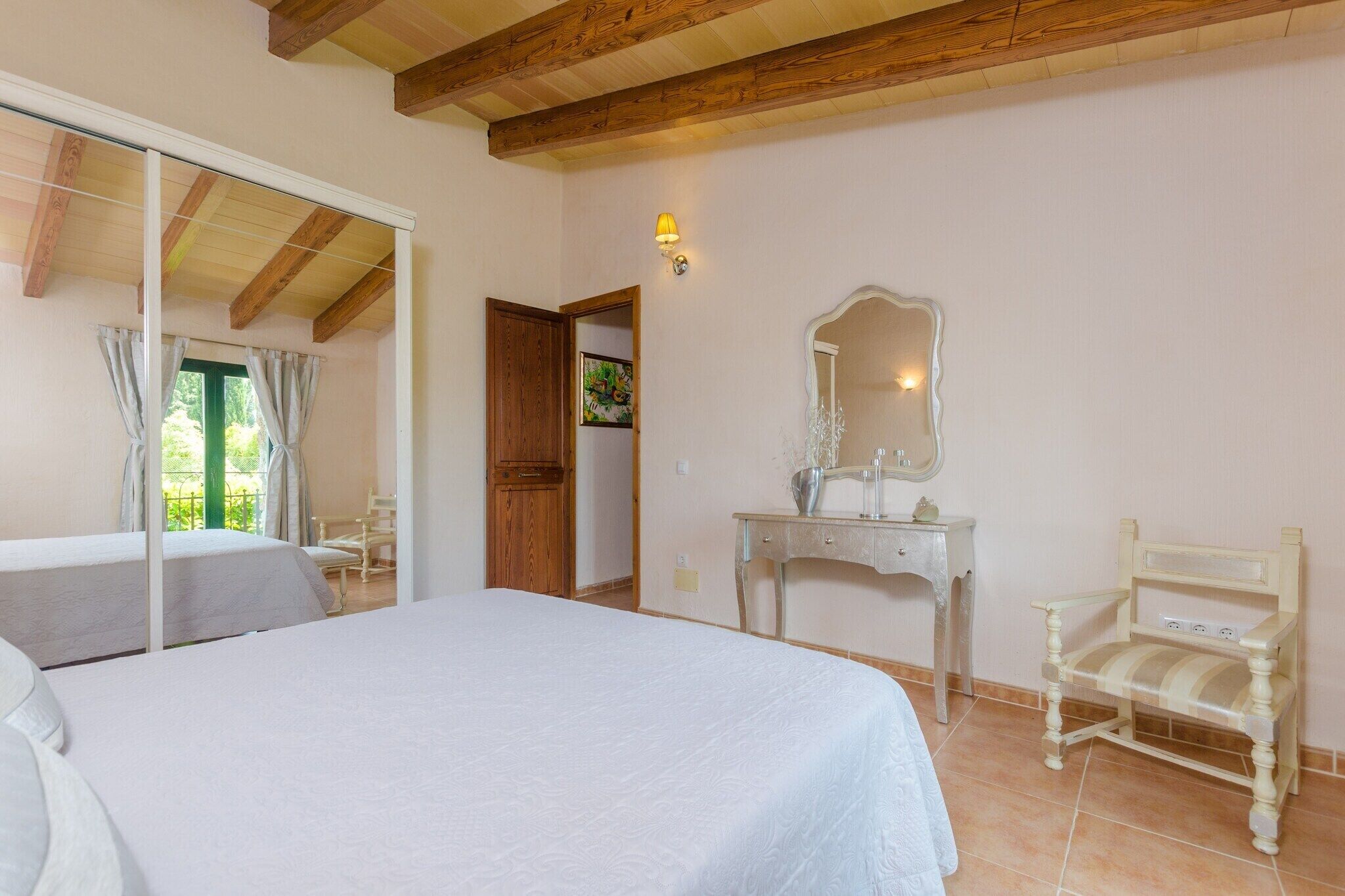 Un dormitorio con una cama, una silla de madera, un tocador con espejo y una puerta que da a otra habitación.
