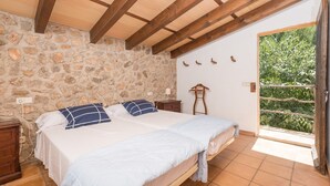 Une chambre avec un mur en pierre, des poutres en bois, un lit avec une literie bleue et blanche, une table de chevet et une vue sur la verdure par une porte ouverte.