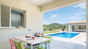A patio with a table set for two, a pool, and a view of a house and hills. - Can Colomer Ferragut  - Villa With Private Pool (Artà, Illes Balears)