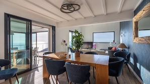 A modern dining area with a wooden table, blue chairs, a potted plant, and a large window offering a view. - Villa Vue Mer 180 - Face aux îles d’Or (Carqueiranne)