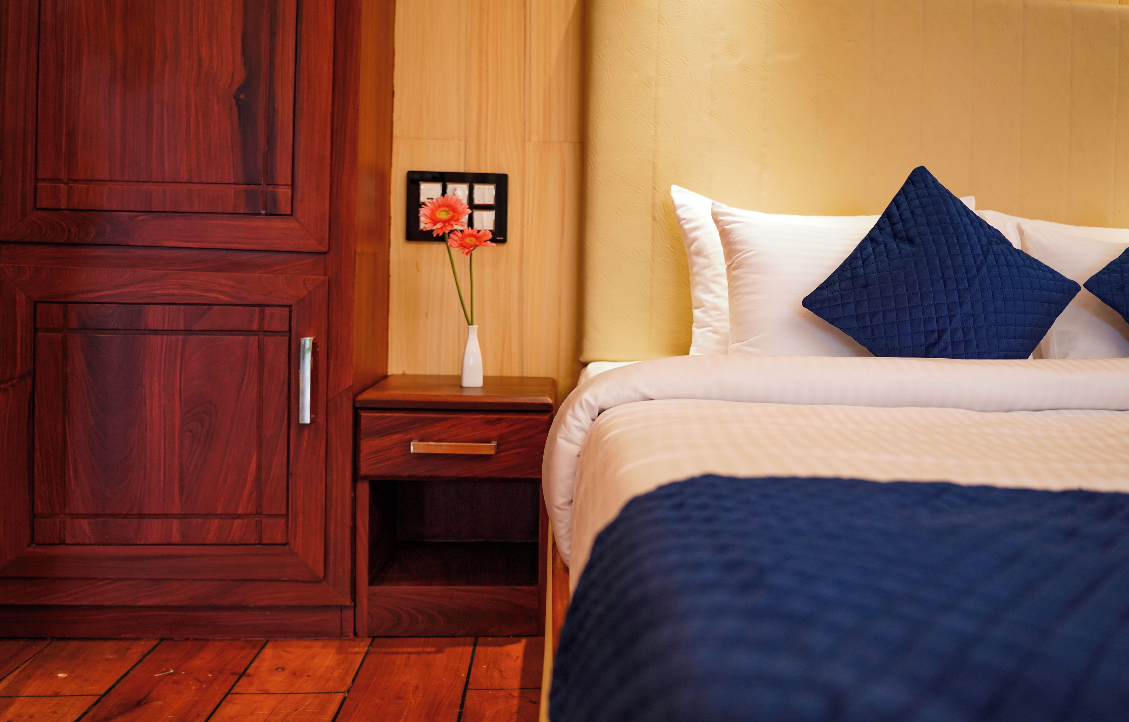 A hotel room with a wooden wardrobe, a bedside table with a vase of flowers, and a bed with white and blue bedding.