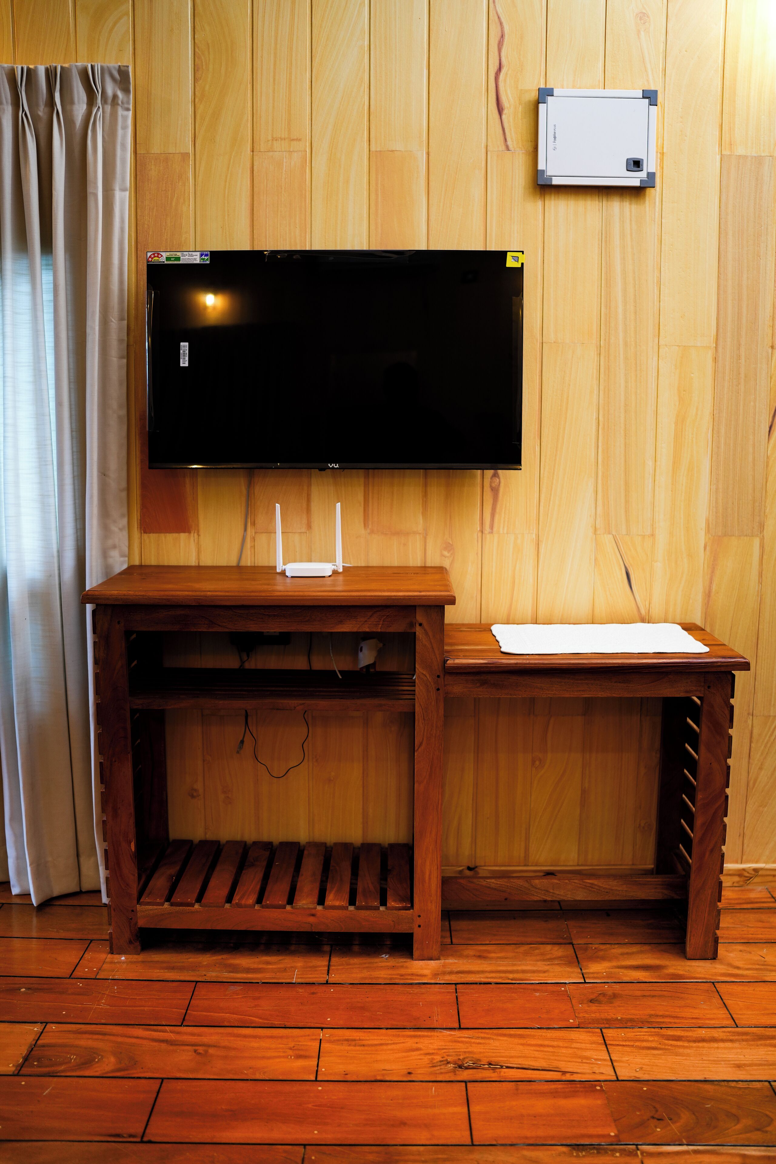 Un mueble de madera para entretenimiento con un televisor de pantalla plana, un pequeño dispositivo blanco en la pared y un banco de madera.