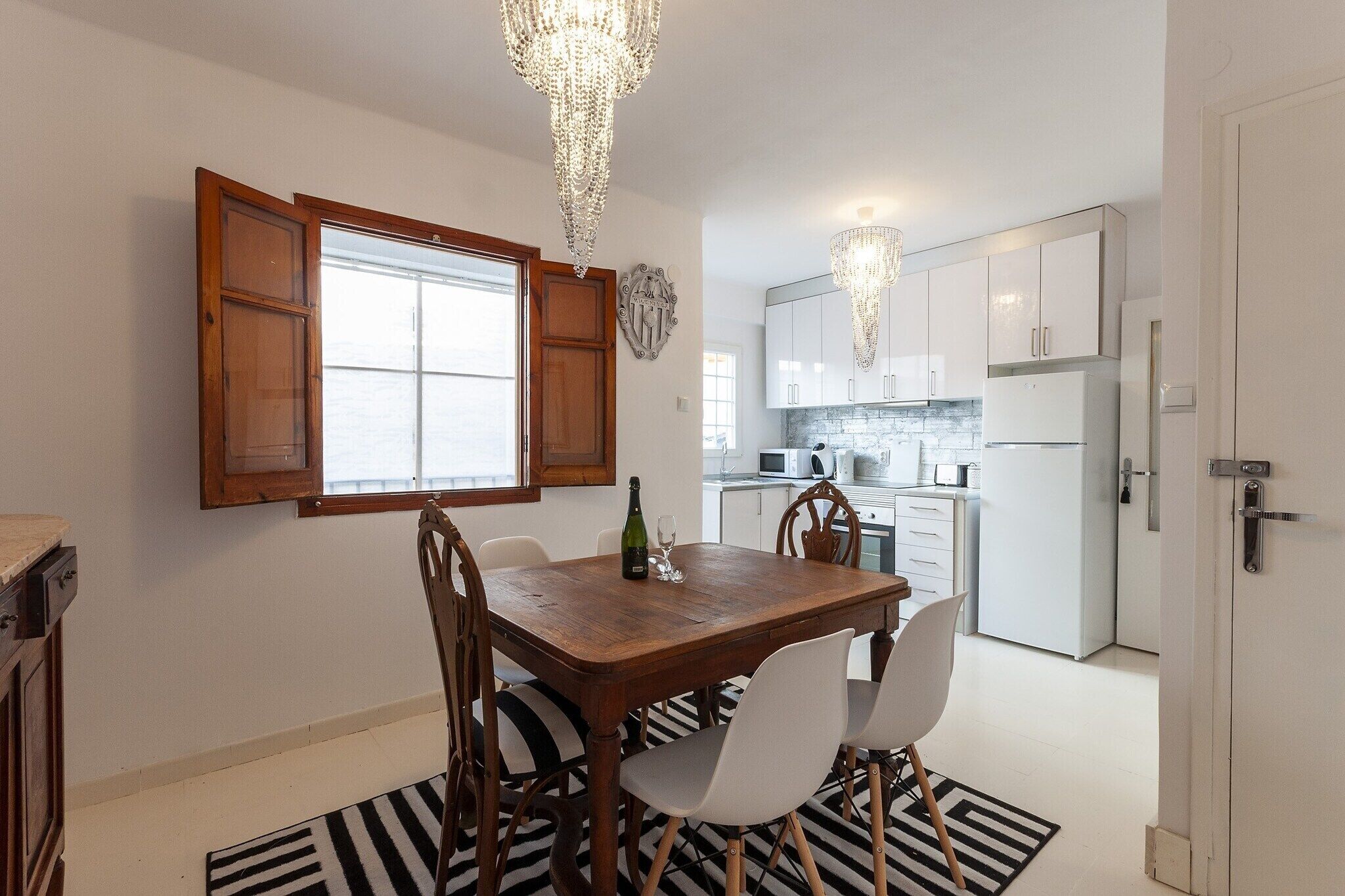 A dining area with a wooden table and chairs, a chandelier, and a view of the kitchen in the background.