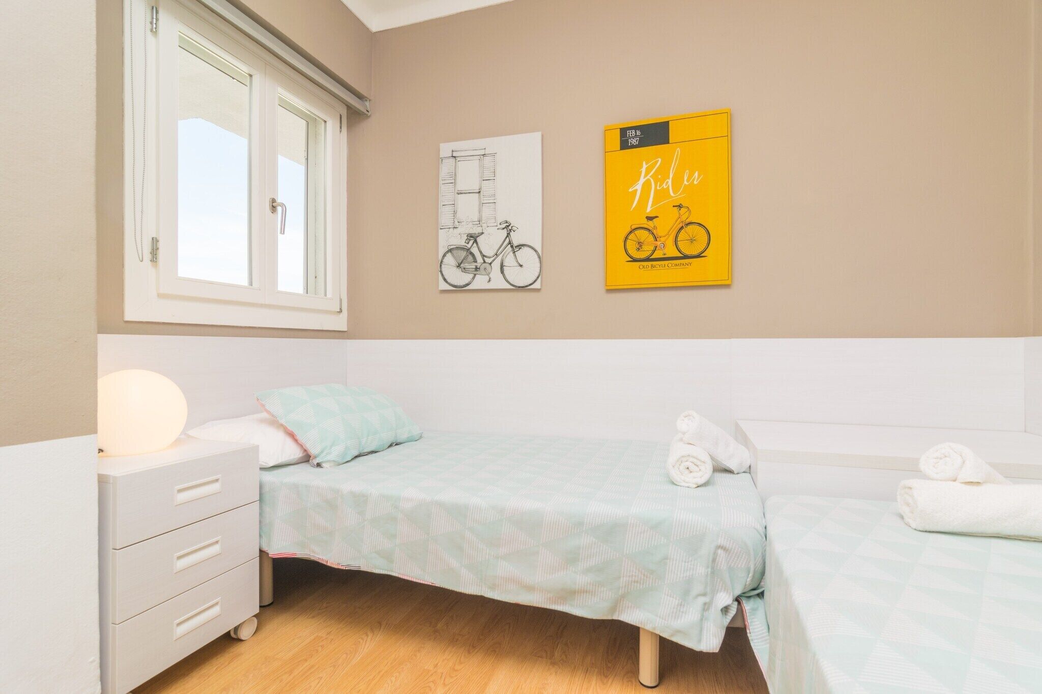 A bedroom with two beds, a window, a nightstand, and wall art.