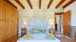 A bedroom with a stone wall, wooden wardrobe, two bedside tables with lamps, a bed with colorful bedding, and a small wooden door. - Can Sua - Villa With Private Pool In Artà (Artà, Illes Balears)