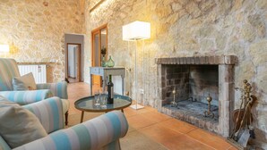 Un salon chaleureux avec un mur en pierre, une cheminée, un fauteuil à rayures, une table ronde avec une bouteille et des verres, et un lampadaire.