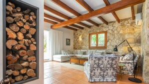 Un salon avec un mur en pierre, des poutres apparentes, une cheminée avec des bûches, un canapé et un fauteuil à motifs.