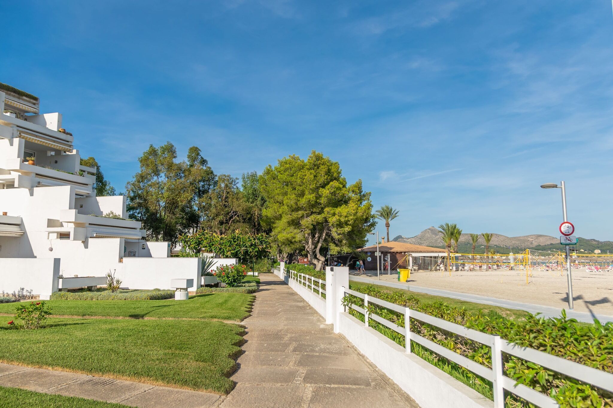 A pathway leading to a beach area with a white fence, greenery, and a clear blue sky.