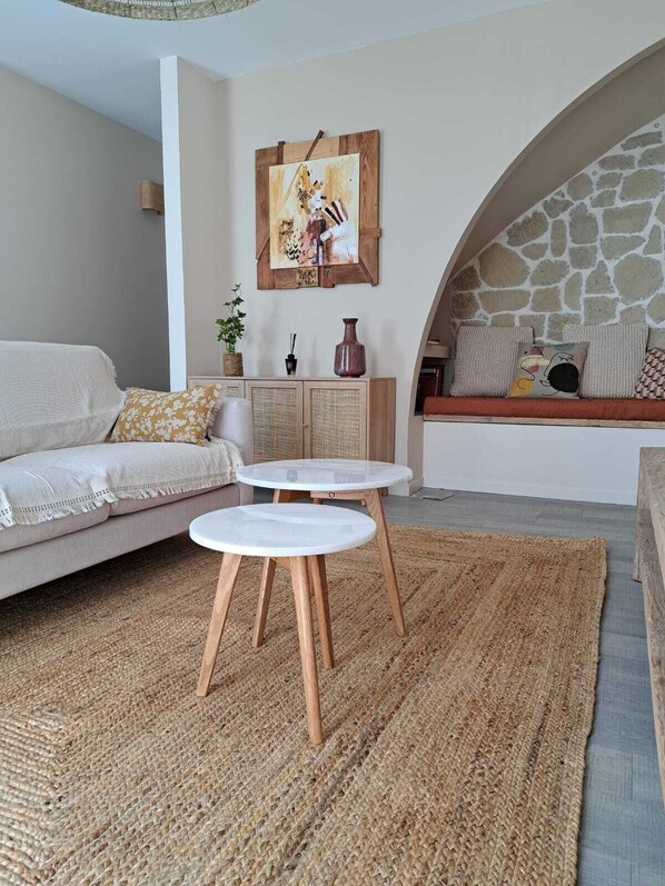 A cozy living room with a sofa, two white round tables, a woven rug, a stone wall, and a painting on the wall.