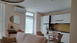 A modern living room with a sofa, a dining table with chairs, a kitchen area with white cabinets, and a large window with blinds. - Beautiful renovated village house in the heart of Provence. (Graveson)