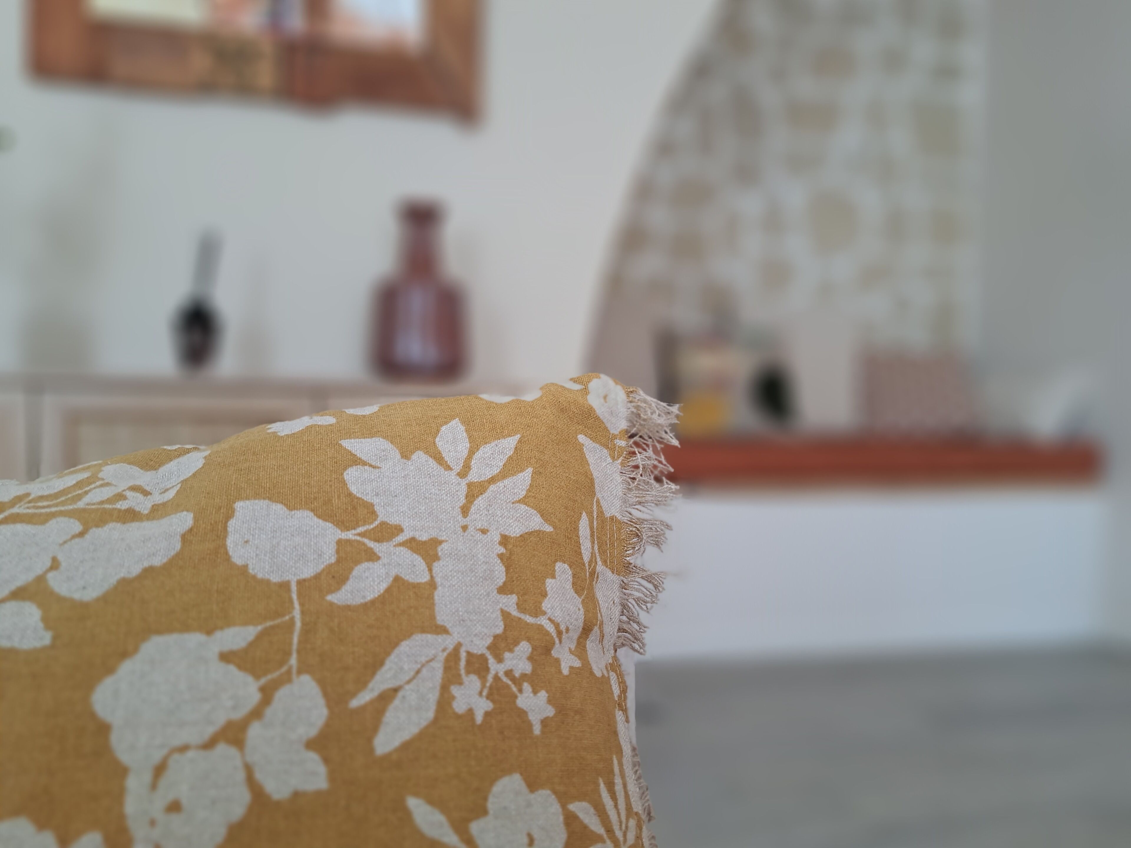 A close-up of a floral-patterned cushion with a frayed edge, set against a blurred background of a room with a wooden cabinet and a mirror.