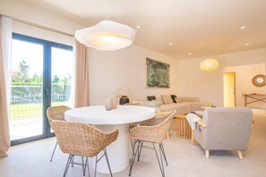 A modern living room with a round dining table, wicker chairs, a grey sofa, and a large window with a view of greenery.