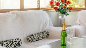 A room with a white sofa, a patterned pillow, a bottle of champagne, two glasses, and a vase with flowers.