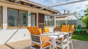An outdoor dining area with a table set for two, surrounded by white chairs with orange cushions.