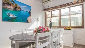 A dining area with a round table, white chairs, a painting of a coastal scene, a window with a view, and a small cabinet with fruit.