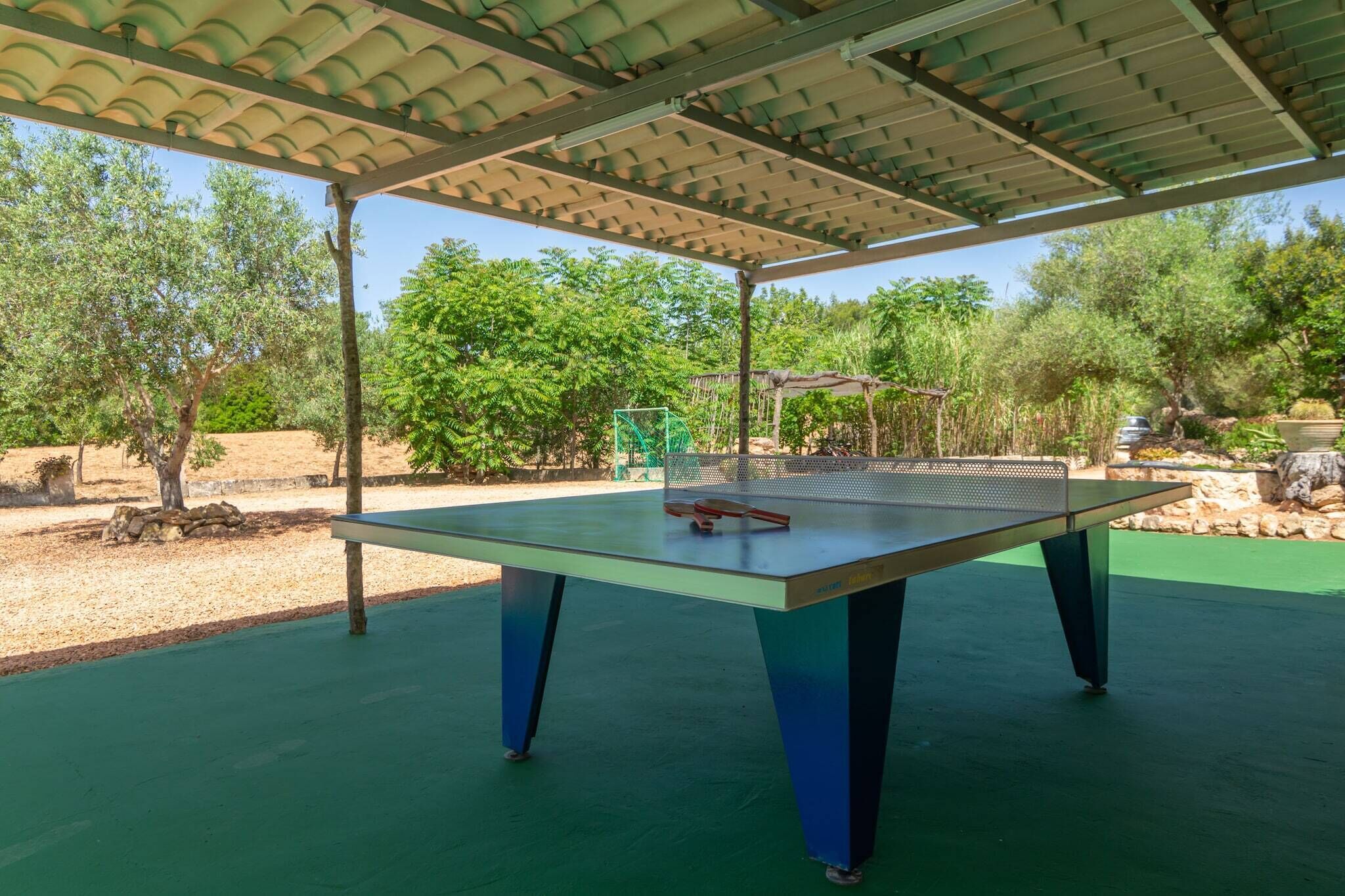 Mesa de ping pong al aire libre bajo una pérgola, con piso verde, rodeada de árboles y rocas.