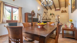 A dining area with a wooden table, chairs, and a fruit bowl. There is a chandelier, a stone wall, and a fireplace.