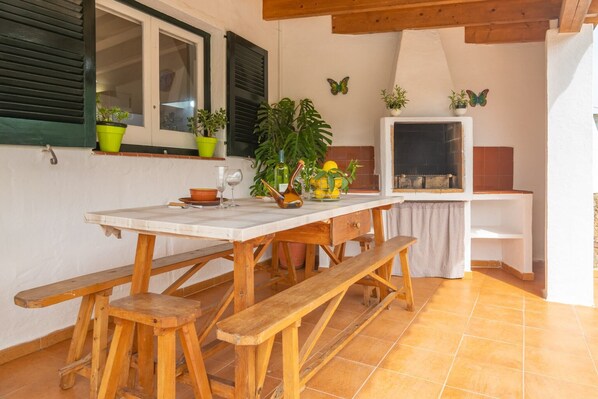 A wooden table with benches, potted plants, and a fireplace in a room with tiled flooring and wooden beams.